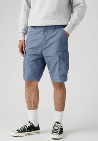 Man wearing blue cargo shorts, white crew socks, black and white sneakers, with hands in pockets, standing against plain background.