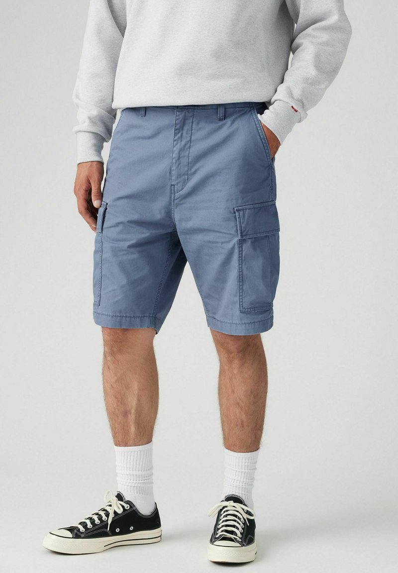 Man med blå cargoshorts, vita strumpor, svartvita sneakers, med händerna i fickorna, som står mot en enfärgad bakgrund.