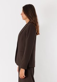 Blazer marrone su misura con rever a notched, spalle strutturate, due tasche e una texture del tessuto liscia. Indossato con pantaloni abbinati.