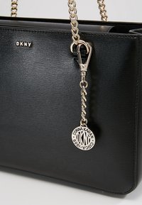 Sac à main en cuir texturé noir de DKNY avec des anses en chaîne dorée et un charme rond en or gravé avec le nom de Donna Karan et le logo.