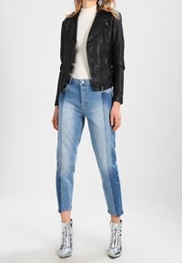 Černá kožená motorkářská bunda přes bílou svetr, doplněná světlými modrými džíny a kovově stříbrnými kotníkovými botami.