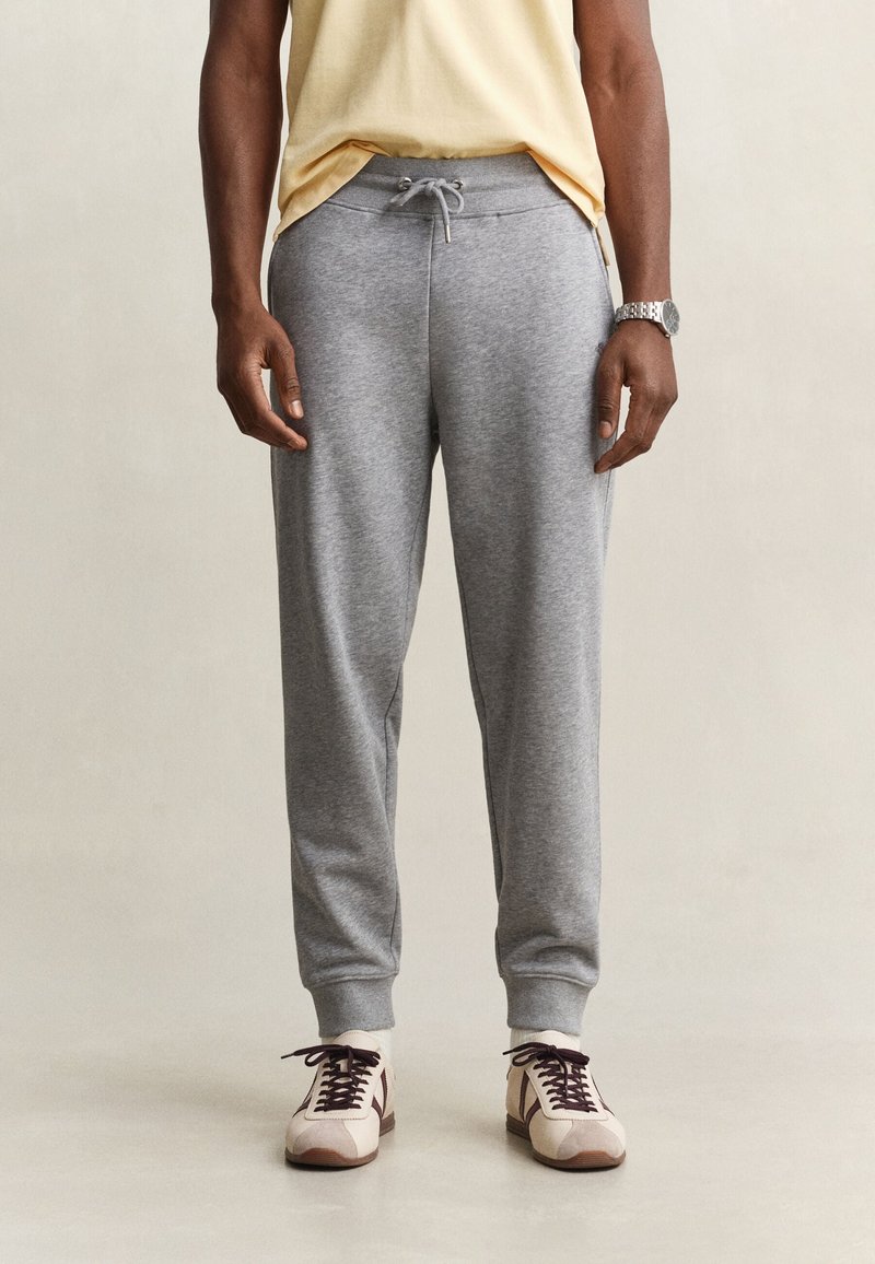 Un homme porte un pantalon de jogging gris clair avec une taille à cordon, des baskets beige et marron, et une chemise jaune, debout devant un fond uni.
