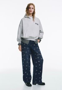 Felpa grigia oversize con tasca frontale, abbinata a pantaloni in denim scuro a gamba larga con dettagli ad anelli circolari. Scarpe nere completano il look.