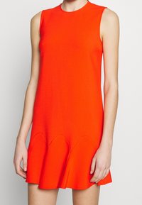 Červené šaty bez rukávů s přiléhavým korzetem, rozšířeným lemem a vroubkovanými detaily na spodní části, vyrobené z hladkého, elastického materiálu.