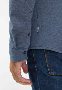 Blaue Herringbone-Hemd mit Knöpfen und langen Ärmeln in Blau, kontrastierenden Knöpfen und einem strukturierten Stoff, kombiniert mit dunklen Jeans.