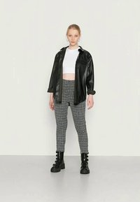 Camisa preta em imitação de couro, top cropped branco, calças de cintura alta xadrez e botas de combate pretas; modelo está contra um fundo neutro.