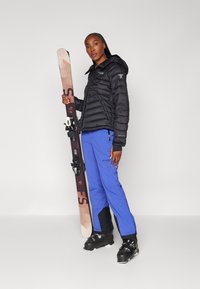 Svart jakke med fyll, blå ski bukser, holder brune ski. Har glidelåser, isolert design og skistøvler med svarte detaljer.
