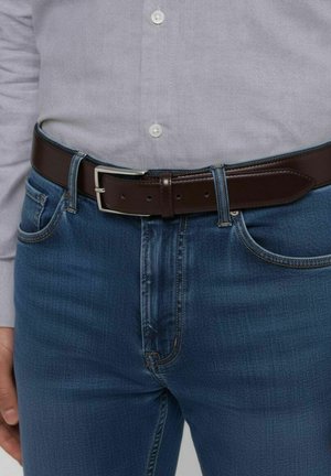 Brauner Ledergürtel mit einer silbernen Schnalle, getragen über blauen Denim-Jeans. Das Hemd ist hellgrau mit Knöpfen und zeigt ein lässiges Outfit.