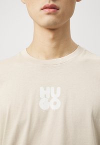 Beige short-sleeve t-shirt made of soft cotton, featuring a prominent white "HUGO" logo with a dripping effect on the front.