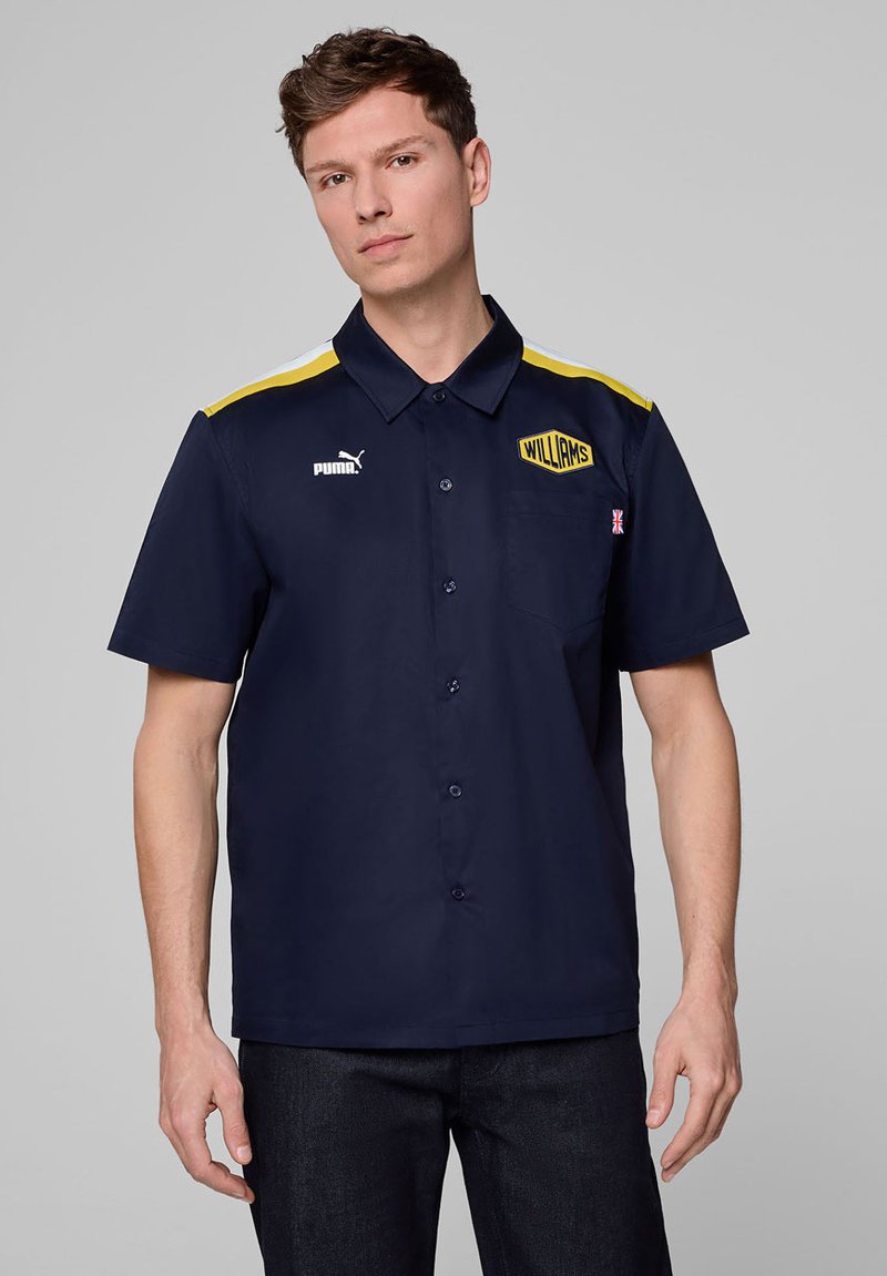 Granatowa koszula krótko rękawowa z guzikami, z żółtymi akcentami i logo Puma. Posiada kieszeń na piersi z logo "WILLIAMS" oraz teksturowany materiał.