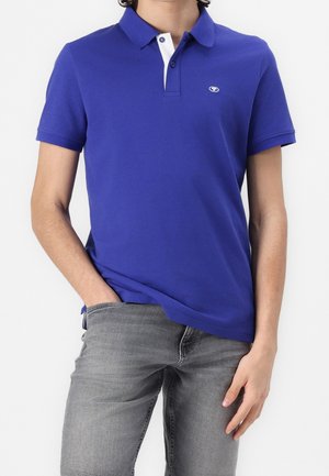 Personne portant un polo bleu à manches courtes avec une patte intérieure blanche et un jean gris, une main dans la poche, face à l'avant.
