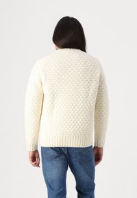 Cream knit sweater with a textured diamond pattern, ribbed cuffs, and hem, paired with blue denim jeans, shown from the back.