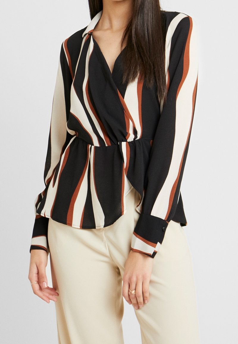 Femme portant un chemisier cache-cœur à manches longues avec des rayures verticales noires, blanches et marron et un pantalon beige clair, debout devant un fond uni.