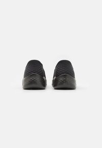 Svarta slip-in skor med en texturerad mesh-oversida, rund tå och diamantmönster på gummisulan, med en slät svart bakdel.