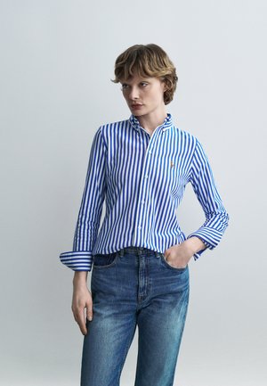 Persona vistiendo una camisa abotonada de rayas verticales azules y blancas con las mangas remangadas y jeans azules, de pie con una mano en el bolsillo.