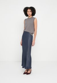 Top gri fără mâneci, tricotat, combinat cu o fustă vaporoasă din satin bleumarin. Model purtând pantofi burgundy cu vârf ascuțit, stând pe un fundal alb.