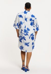 Weißes Kleid mit großem blauen Blumenmuster, kurzen Ärmeln und einem Kragen. Kombiniert mit blauen Sandalen mit dicker Ferse. Verfügt über eine lockere Passform.