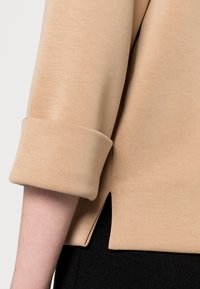Beige toppi, jossa on kääritty hihansuu ja sivuhalkiot. Kangas on sileä ja siinä on hieno tekstuuri. Mustaa materiaalia näkyy alla.
