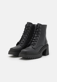 Bottines noires à talon épais, à lacets sur l'avant, avec fermetures éclair latérales et semelles épaisses à crampons sur fond blanc uni.