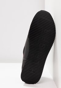 Semelle de chaussure de sport noire avec des rainures horizontales et un logo de marque en relief, photographiée sur un fond d'angle blanc.