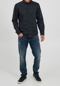 Camisa oscura abotonada con cuello, combinada con vaqueros azul claro y zapatillas negras. La camisa presenta una textura sutil y detalles con botones.