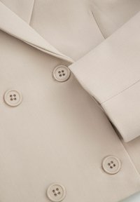 Podwójnie zapinany płaszcz w jasnobeżowym materiale, charakteryzujący się gładką teksturą, wyraźnymi klapami i czterema okrągłymi guzikami.