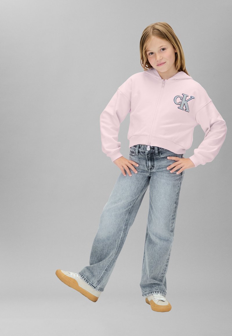 Sudadera rosa recortada con cremallera y logo "CK", combinada con jeans acampanados azul claro y zapatillas crema con suela texturizada.