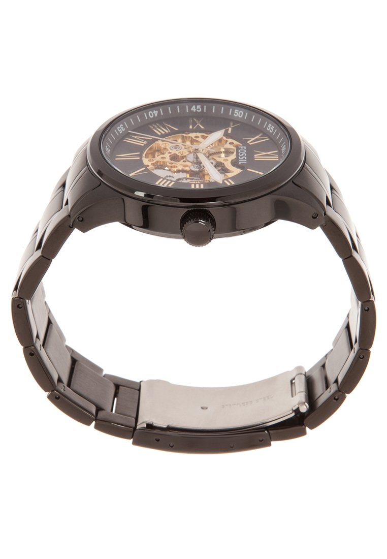 Reloj de metal negro con un dial esqueleto, números romanos y una correa texturizada. Presenta una caja redonda y una corona en el lateral.