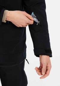 Zwarte jas met een met ritssluiting afgesloten mouwzak. Hand die een item invoegt. Klittenband voor aanpassing van de manchetten. Materiaal lijkt textuur en duurzaam te zijn.