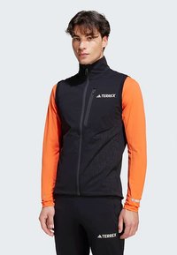 Schwarze ärmellose Weste mit hohem Kragen, ausgestattet mit einem Reißverschluss und einem gemusterten Seitenbereich. Kombiniert mit einer orangefarbenen Langarm-Basisschicht.