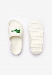 Sandalias deslizantes blancas hechas de goma con una suela texturizada, que presentan un logo de cocodrilo verde en la correa. Diseño plano con punta abierta.