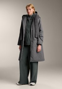 Vadderad grå kappa med huva, prydd med en texturerad mörkgrå scarf. Bärs över en mörkgrön outfit, som kompletterar vida byxor och sneakers.