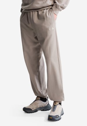 Personne portant un pantalon de jogging taupe avec le logo The North Face et des chaussures de randonnée beiges avec semelles noires et marquage Vibram.
