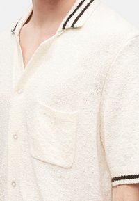 Gros plan d'une chemise à manches courtes texturée crème avec col et poignets rayés foncés, devant boutonnée, et poche poitrine.
