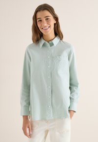 Jeune femme souriante, portant une chemise à rayures vert clair et blanches boutonnée, associée à un pantalon blanc, se tenant devant un fond beige uni.