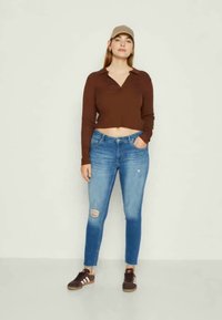 Hnědý krátký top s dlouhým rukávem, modré skinny džíny s opotřebovanými detaily a hnědé tenisky s bílými pruhy, které nosí stojící osoba.