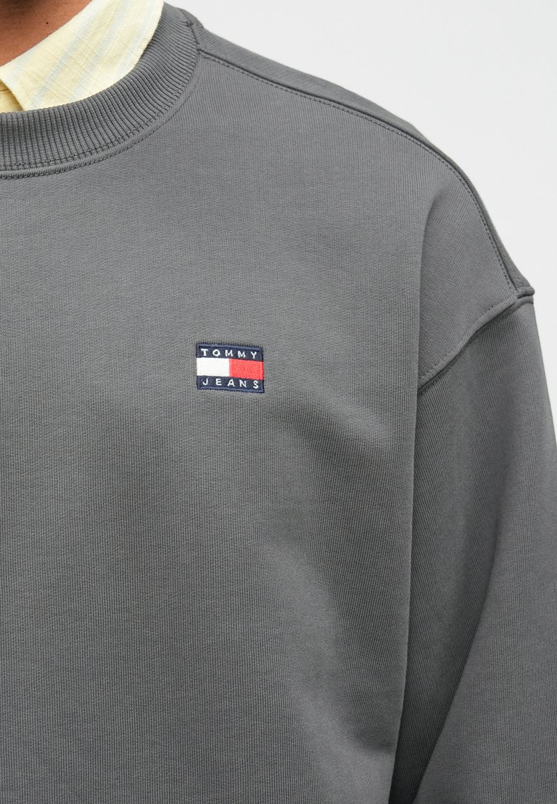 Gray sweatshirt with ribbed crew neckline, featuring a small blue and red "TOMMY JEANS" logo patch on the chest. Smooth texture, casual design.
