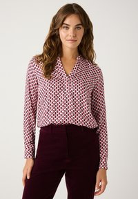 Langarmbluse in Weiß mit einem roten und marineblauen kreisförmigen Muster, mit eingekerbtem Kragen und Knopfblende, kombiniert mit bordeauxfarbener Cordhosen.