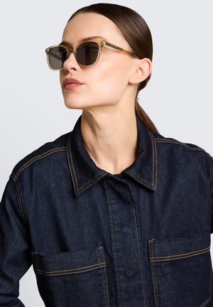 Femme aux cheveux châtain foncé en queue de cheval basse, portant des lunettes de soleil brunes translucides et une veste en jean foncée avec des coutures orange.