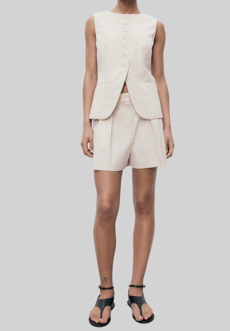 Femme portant un gilet beige sans manches boutonné, un short taille haute assorti et des sandales noires à bout ouvert avec des brides à la cheville.