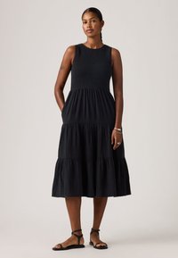 Γυναίκα που στέκεται, ντυμένη με μαύρο αμάνικο φόρεμα με στρώσεις μέχρι το μέσο της γάμπας και μαύρα σανδάλια με λουράκι στον αστράγαλο, μπροστά σε απλό φωτεινό φόντο.