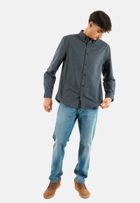 Camisa de botones en tela gris oscuro, de manga larga, con cuello clásico. Combinada con jeans azules claros y zapatos marrones, en posición de pie.