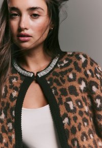 Leopardikuvioinen neuletakki pehmeässä ruskeassa ja mustassa neuloksessa, helmikoristeltu kaulus ja pyöreä pääntie, teksturoitu viimeistely.