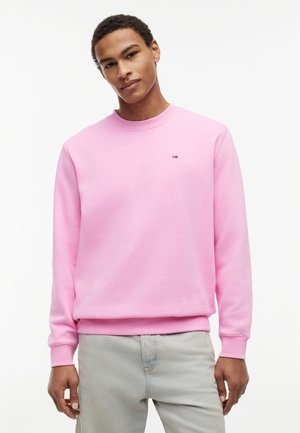 Joven con sudadera rosa claro con pequeño logo en el pecho y pantalones gris claro, de pie contra un fondo blanco liso.