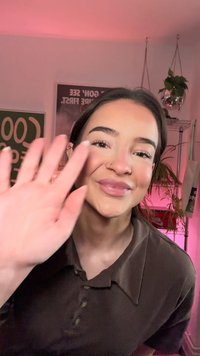 Mlada ženska s temnimi lasmi in nosnim obročkom se smehlja in maše v sobi z rožnato osvetlitvijo, v ozadju pa so viseče rastline in plakati.