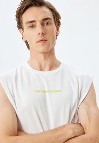 Witte mouwloze T-shirt, gemaakt van structuurstof, met een groene print die "KEEP MOVING FORWARD" over de borst leest.