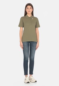 Polo shirt verde oliva con colletto e tre bottoni, abbinato a jeans skinny blu e sneakers chiare con accenti più scuri.