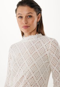 Top in pizzo bianco con collo alto, caratterizzato da un motivo a diamante e una finitura testurizzata. Maniche lunghe e bordi delicati a scallope.