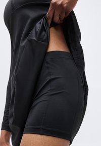 Άτομο σηκώνει μαύρο εξωτερικό σορτς για να αποκαλύψει μαύρο εφαρμοστό εσωτερικό σορτς που φοράει από κάτω.