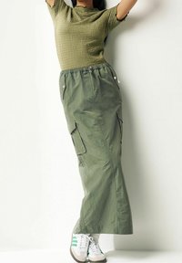 Frau trägt ein olivgrünes, strukturiertes, kurzärmliges Oberteil, einen passenden langen Cargo-Rock und weiße Sneaker mit grünen Streifen und posiert mit erhobenen Armen.